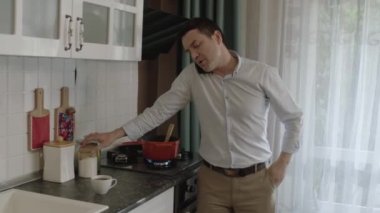 Young single man preparing food in kitchen and talking on cell phone at the same time. The man who lives alone follows his work with his smartphone while he prepares food for himself.