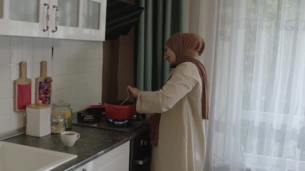ヒジャーブ州の若いイスラム教徒の女性は台所で食べ物を準備し 同時に携帯電話で話しています 自分のために食べ物を準備しながら 彼女のスマートフォンで話す女性 — ストック動画