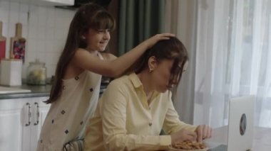 Young mother working remotely with laptop in the kitchen. The little girl hugs her mother from behind, ruffles her hair, shakes her shoulders, tries to tell her mother to take time for herself.