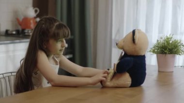 Little girl and teddy bear holding a tablet computer in their laps. Girl doing school homework at kitchen table,tablet computer with green screen, studying online,watching videos,playing online games.