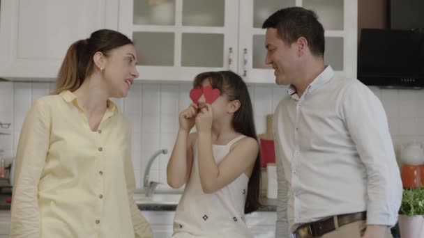 台所で両親と楽しい時間を過ごしながら 目に紙の心を持つ小さな女の子 少女は誕生日 母の日 父の日を目に紙の心を持って祝う — ストック動画