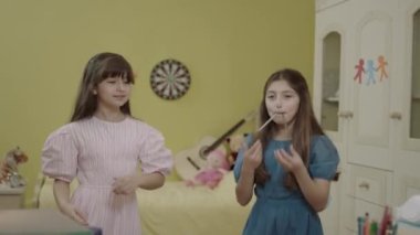 Little girls are having fun together by dancing in their room at home. Happy girls are having a crazy party at their house. Funny kids dancing, jumping, singing are sending air kisses to the camera.