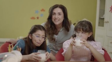 Caring mother brings milk to little girls doing school homework. The caring parent who brings two glasses of milk to their young children to take a break from their lessons. Ideal mother portrait.