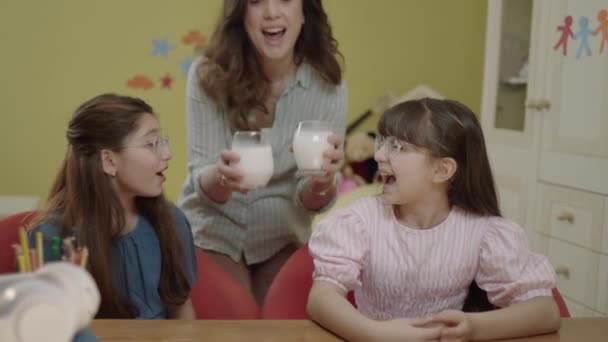 思いやりのある母親は 学校の宿題をしている小さな女の子に牛乳をもたらします 幼い子供たちに2杯の牛乳を持ってくる思いやりのある親は 彼らのレッスンから休憩を取るために 理想的な母像 — ストック動画