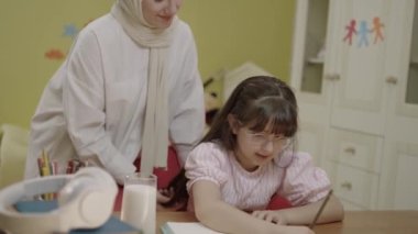 Smart elementary school girl learning to write, doing math homework, sitting at home desk. A loving headscarved mother brings milk for her little girl to drink while she does her schoolwork.