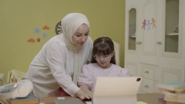 Smart elementary school girl doing math homework with tablet computer, sitting at home desk. Loving hijab-clad mother helps her little daughter solve problems while doing her homework.