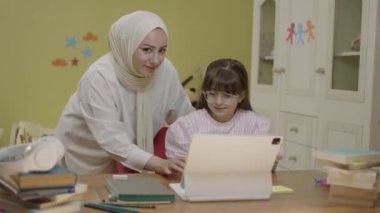 Smart elementary school girl doing math homework with tablet computer, sitting at home desk. Loving hijab-clad mother helps her little daughter solve problems while doing her homework.