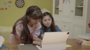 Smart elementary school girls doing math homework with tablet computer, sitting at home desk. Little girls succeed by solving questions while doing their homework.