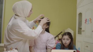 Smart elementary school girls doing math homework with tablet computer, sitting at home desk. Loving hijab mom helps little girls solve problems while doing their homework. Ideal mother portrait.