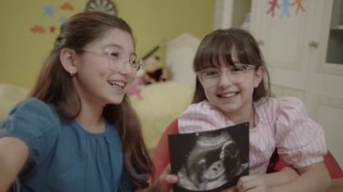 Little girls holding an image of their unborn sibling from ultrasound. Girls take selfie with ultrasound photo of their pregnant mother, describe surprise on video call.