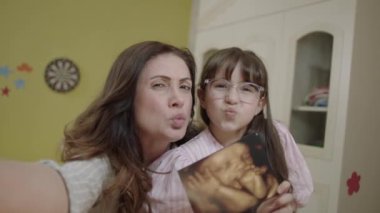 Little girl holding an image of her unborn siblings from ultrasound. Girl takes selfie with ultrasound photo of pregnant mother, describes surprise on video call. Happy family portrait.