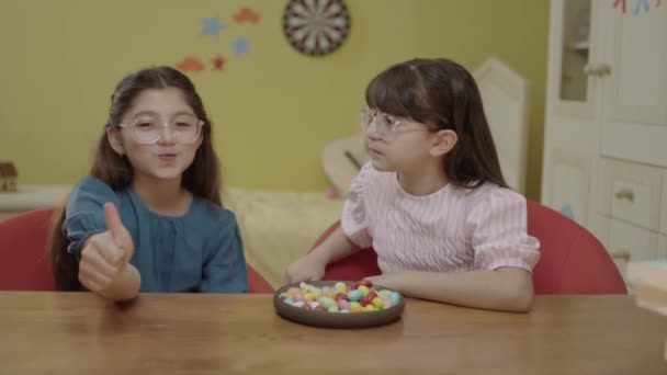 小さな女の子は学校の宿題のクラスから休憩を取り カラフルなキャンディーを食べる 小さなガールフレンドは彼女の部屋でテーブルの上にカラフルなキャンディーを食べ 笑って一緒に楽しみます — ストック動画
