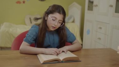 A little girl with long hair is reading a book in her room. Cute little girl reading a book, turning the page, learning, working, studying, reading a book.