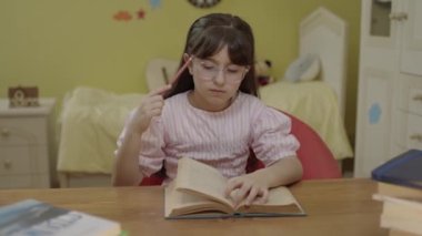 A little girl with long hair is reading a book in her room.Cute little girl reading a book, turning the page, learning, working, reading, reading a book. Portrait of little girl doing school homework.