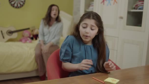 在家里 小女孩坐在桌旁 用彩色的小纸片列着要做的事 年轻的母亲警告女儿读书和做作业 这个女孩不想读书或读书 — 图库视频影像