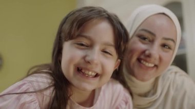 Little cute girl taking selfie with her young mother in hijab, recording video, photographing beautiful moment. Mother looking at camera while daughter enjoying video call. Front phone camera view.