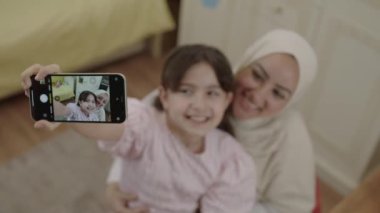 Little cute girl taking selfie with her young mother in hijab, recording video, photographing beautiful moment. Mother looking at camera while daughter enjoying video call. Front phone camera view.