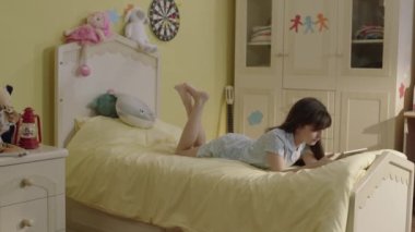 Little girl surfing internet with tablet computer lying on her bed at home.Cute girl continues distance education on tablet in bedroom. She watches funny videos and plays online games she.