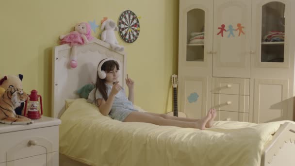 小女孩一个人在床上唱歌 带着耳机弹奏着想象中的吉他 女孩在家里的房间里用一个假想的话筒 把自己想象成一个真正的歌手 — 图库视频影像