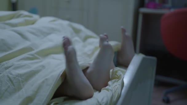 楽しそうに笑っている子供たちの足はキルトの下から出てきます 子供たちはベッドの上に裸足で横たわって足を動かす 居心地の良い家の朝 目覚め — ストック動画