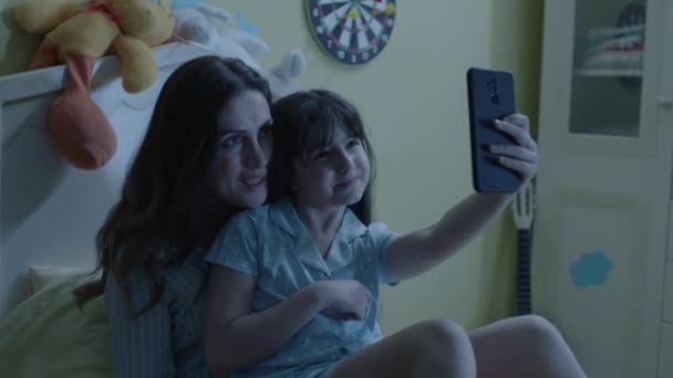 幸せな母親の小さな娘は スマートフォンで夜の視聴ビデオで快適なベッドに横たわって 写真を見て 自撮りのポーズ 電話でのビデオ通話 遠隔会話を楽しんでいます — ストック動画