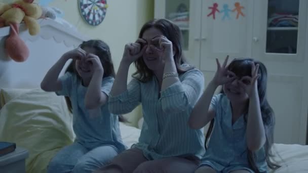 彼らの小さな娘と幸せな若い母親は週末に楽しんでいます 楽しいと面白い家族は夜にベッドに座って 双眼鏡を指で作る — ストック動画