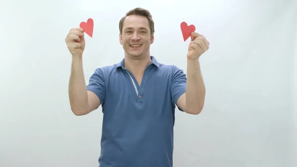 Happy young man holding two small paper hearts in hands isolated on white background. Love in your eyes, romantic concept. Video of a smiling man holding two paper hearts.
