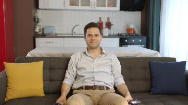 Young man sitting on comfortable sofa in his apartment and looking at camera with smile. Creative people can put whatever they want where the man points.