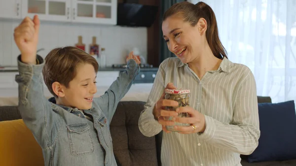 stock image Young mother and her little son putting coins in piggy bank while sitting on sofa at home. The mother teaches the child to be thrifty, to save money, to think about the future, to save.