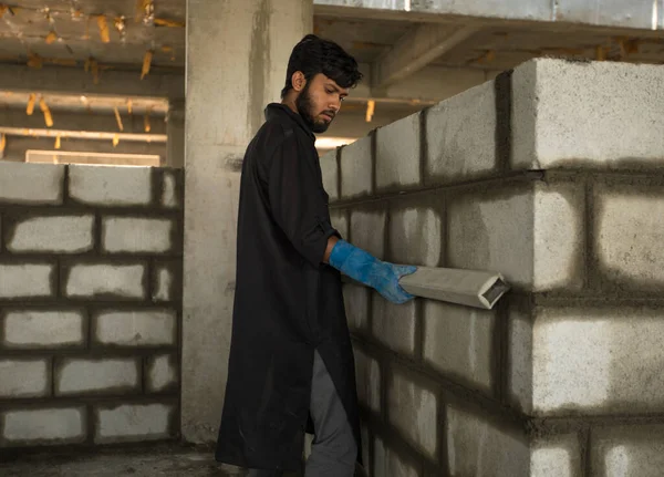 Bir işçi tuğla duvarı incelemek için bir seviye kullanırken görülebilir. İşçi, iş kıyafetleri ve eldiven giyerken duvarın düzgün olduğundan emin olmaya çalışıyor.. 