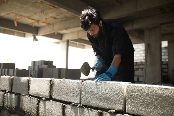 Tuğla ve çimento kullanan bir inşaat işçisi. İşçi elindeki işe yoğunlaşır ve iş için iş kıyafeti ve eldiven giyer..
