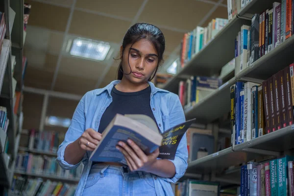 Üniversite kütüphanesinde kitap arayan ve eğitim, öğrenim ve araştırma için okul, üniversite ya da üniversite kampüsünde doğru kitabı seçmek üzere kitap okuyan çekici bir kız..