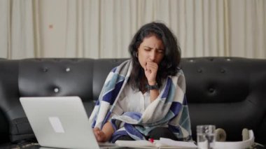 Kanepede oturan sıcak ekoseyle kaplı hasta kadın iş yerinde grip, buruşmuş peçeteler semptomları hafifletmek için ilaçlar, dizüstü bilgisayardan alınan belgelerle çalışıyor. Mevsimlik grip konsepti