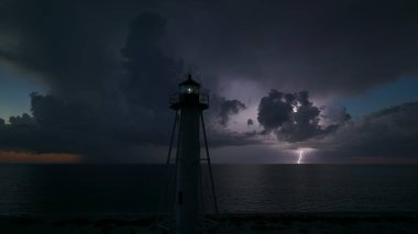 Deniz kıyısındaki uzun deniz feneri fırtınalı bir gecede ticari gemilerin seyrüsefer için yanıp sönen ışık. Okyanus üzerinde şimşekler çakan fırtına gemiler için tehlike arz ediyor..