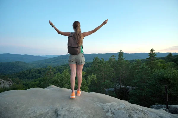 Hiker woman standing on mountain footpath raising up hands enjoying evening nature during travel on wilderness trail. Lonely female traveler traversing high hilltop route. Healthy lifestyle concept.