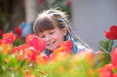 Mutlu kız çocuğu yaz bahçesinde oynuyor güneşli bir günde kırmızı lale çiçeklerinin tatlı kokusunun tadını çıkarıyor..