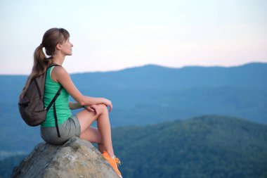 Sportswoman sitting alone taking a break on hillside trail. Female hiker enjoying view of evening nature from rocky cliff on wilderness path. Active lifestyle concept.