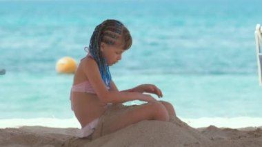 Cute happy child girl playing with sand on ocean beach during summer tropical vacations.