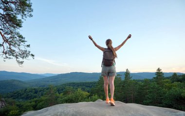 Tepedeki patikada kollarını kaldırmış tek başına duran sportif bir kadın. Dişi yürüyüşçü, vahşi yoldaki kayalık uçurumdan akşam manzarasının keyfini çıkarıyor. Etkin yaşam tarzı kavramı.