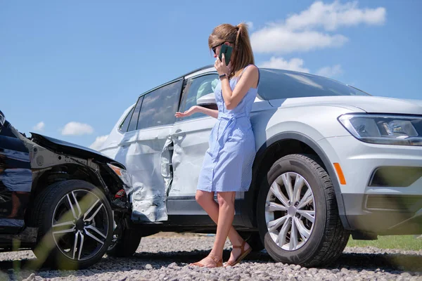 Stressed driver talking on sellphone on roadside near her smashed vehicle calling for emergency service help after car accident. Road safety and insurance concept.