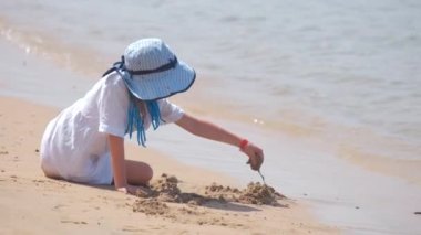 Büyük şapkalı, beyaz elbiseli, mavi gökyüzünün arka planında ıslak kumla oynayan güzel kız çocuğu ve berrak okyanus gölü suyu..