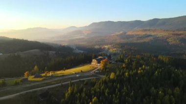 View from above of ukrainian Carpathian mountains with wooded hills and traditional village homes at autumnal sunset. Brightly illuminated pine woods with scattered local settlement houses.