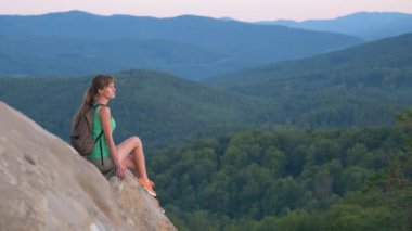 Sportif kadın yamaç yolunda tek başına oturuyor. Dişi yürüyüşçü, vahşi yoldaki kayalık uçurumdan akşam manzarasının keyfini çıkarıyor. Etkin yaşam tarzı kavramı.