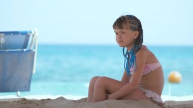 Happy child girl in bikini swimsuit playing with sand on seaside beach during summer tropical vacation.