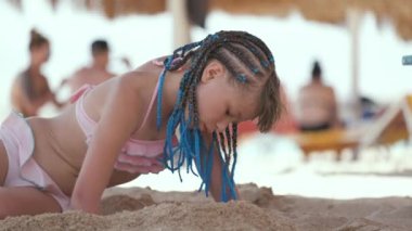Happy child girl in swimsuit playing with sand under umbrella shadow during summer tropical vacations.