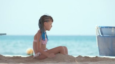 Happy child girl in bikini swimsuit playing with sand on seaside beach during summer tropical vacation.