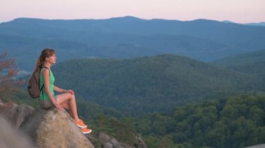 Young woman hiker sitting alone on rocky mountain enjoying view of evening nature on wilderness trail. Active way of life concept.