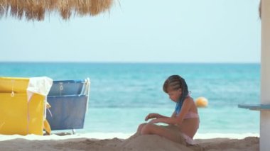 Happy child girl in bikini swimsuit playing with sand on seaside beach during summer tropical vacation.