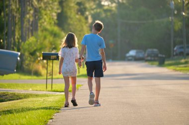 Back view of two young teenage children, girl and boy, brother and sister walking together on suburban street on bright sunny evening.