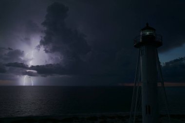 Deniz kıyısındaki uzun deniz feneri fırtınalı bir gecede ticari gemilerin seyrüsefer için yanıp sönen ışık. Okyanus üzerinde şimşekler çakan fırtına gemiler için tehlike arz ediyor..
